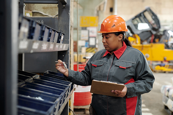 Serious young workwoman in safety helmet and uniform making revision of spare parts for industrial machines in modern plant